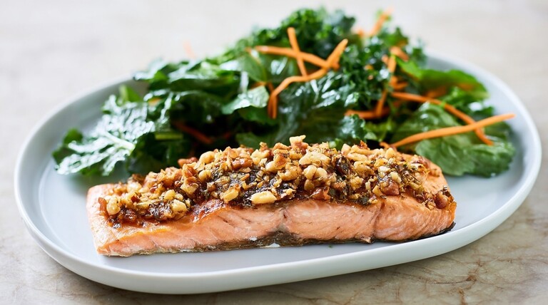 Grilled salmon fillet topped with nuts and herbs on a white plate, served with a fresh green salad and carrot shreds.