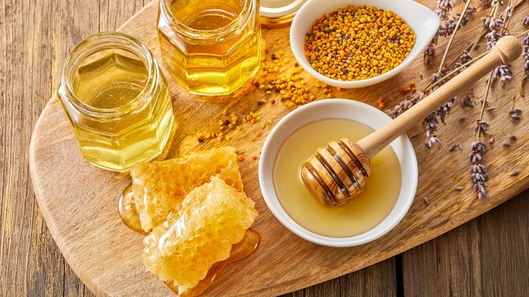 Assorted honey jars, honeycomb, bee pollen, lavender, and a wooden honey dipper arranged on a rustic wooden tray.