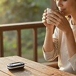 A woman sips from a cup on a sunlit porch, with a glucose meter resting on the wooden table in front of her.