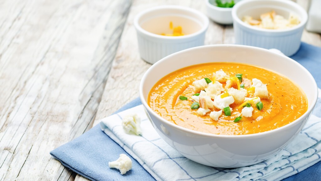 Creamy soup topped with cauliflower, croutons, and green onions in a white bowl on a folded blue napkin.