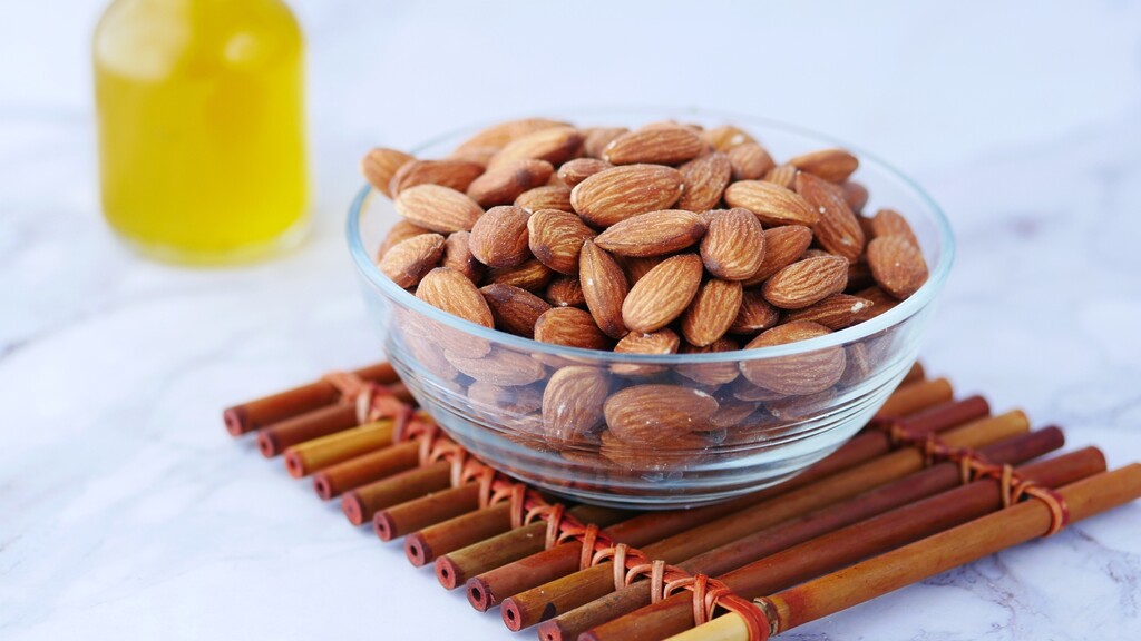 A glass bowl filled with almonds sits on a bamboo mat, with a blurred bottle of oil in the background.