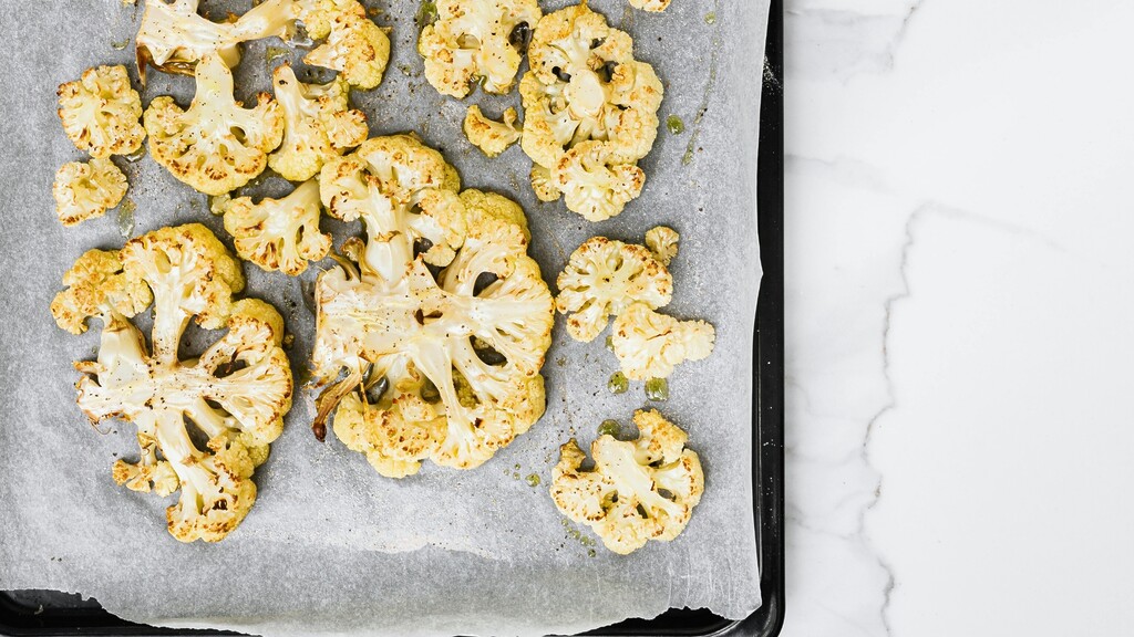 Roasted cauliflower florets spread on parchment paper on a baking sheet beside a marble countertop.