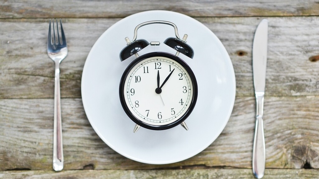 Alarm clock on a white plate between a fork and knife, representing meal timing and intermittent fasting.