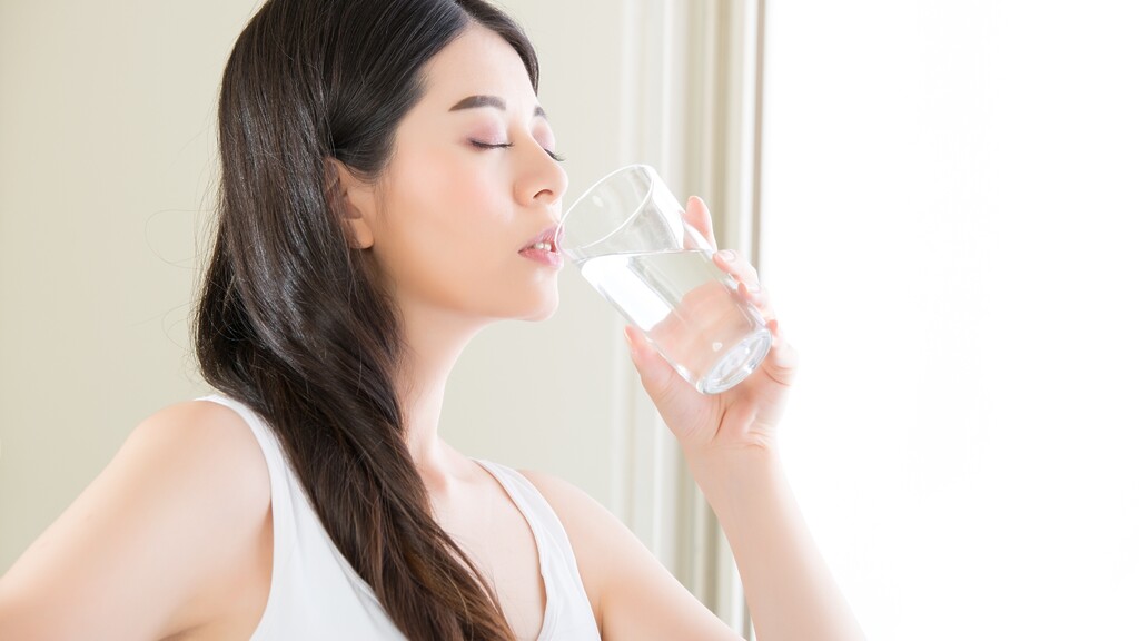 Woman drinking a glass of water in a bright room, suggesting hydration during fasting.