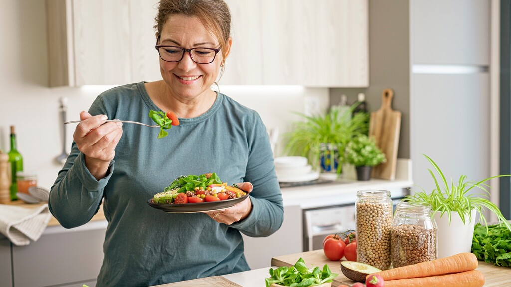 Woman eating a fresh salad in a bright kitchen with vegetables, legumes, avocado, and carrots on the counter.