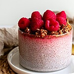 5 Simple Ways to Add Chia Seeds to a Diabetic Diet A glass bowl of pink chia pudding topped with fresh raspberries and granola, served on a white plate with a gold spoon and a mug in the background.