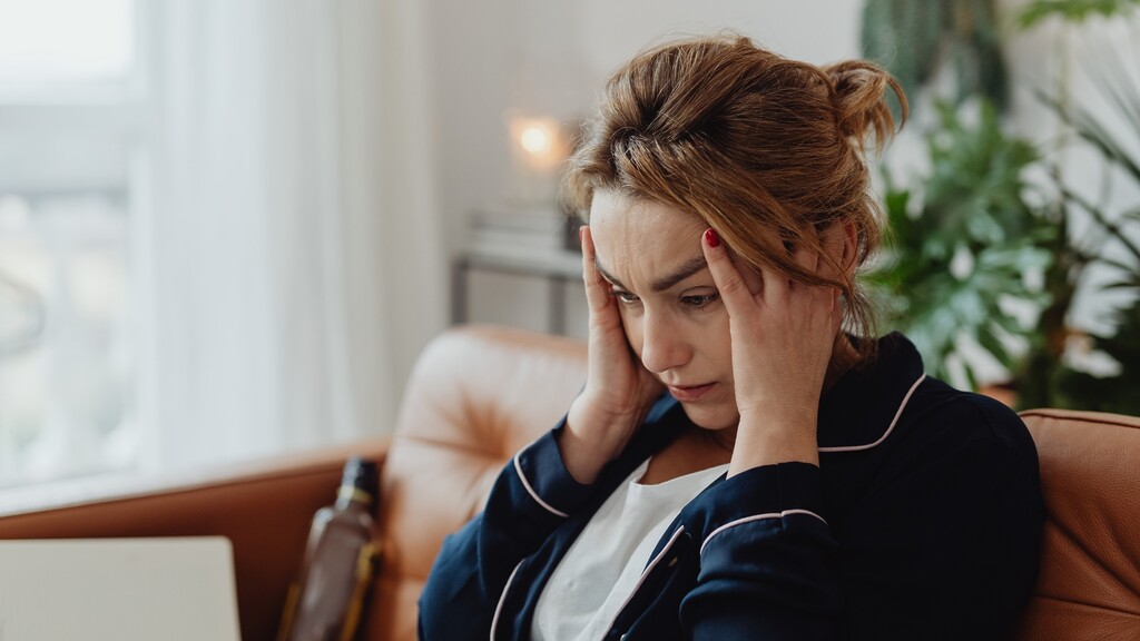 Woman sitting on a couch with her hands on her temples, looking stressed and overwhelmed near a laptop.