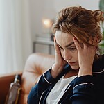 Woman sitting on a couch with her hands on her temples, looking stressed and overwhelmed near a laptop.