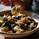 A bowl of pasta with leafy greens and mushrooms, with a person lifting a bite on a fork in warm natural light.