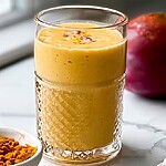 7 Anti-Inflammatory Spices to Add to Your Daily Diet A glass of mango turmeric smoothie on a marble countertop, with a small bowl of turmeric powder, a mango, and coconut pieces beside it.