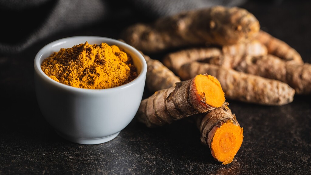 Fresh turmeric root sitting next to a small bowl of bright orange ground turmeric