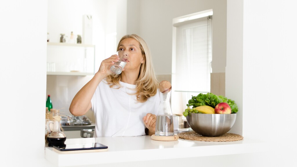 Woman drinking water beside fresh produce in a bright kitchen.