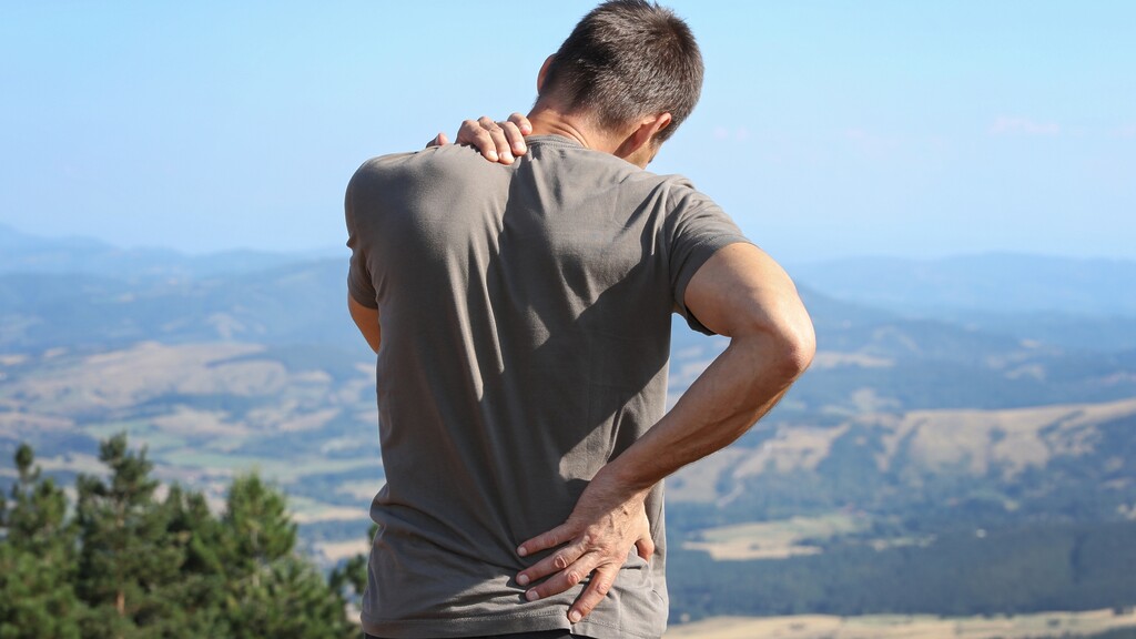 A man stands outdoors with one hand on his lower back and one hand on his shoulder, suggesting back or shoulder pain against a mountain landscape.