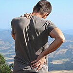 A man stands outdoors with one hand on his lower back and one hand on his shoulder, suggesting back or shoulder pain against a mountain landscape.