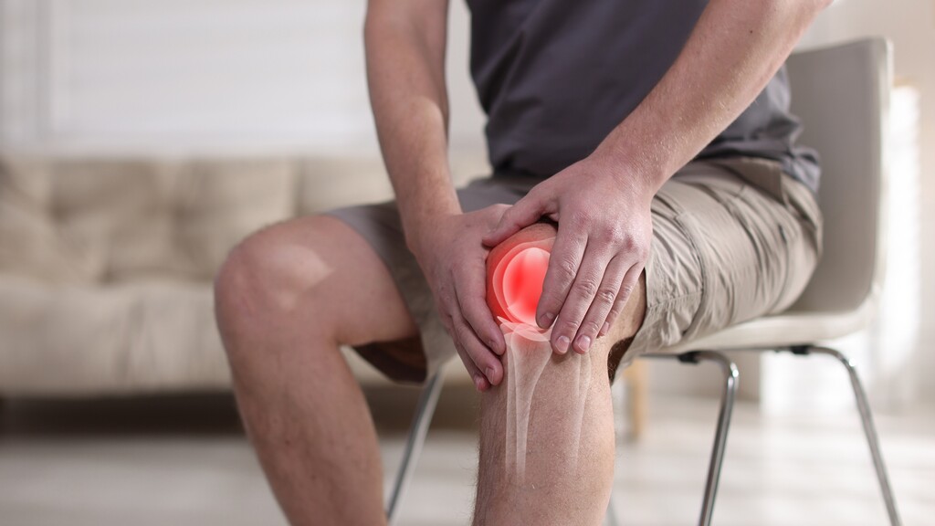 A person sitting on a chair holding their painful knee, with a red highlight showing knee discomfort.