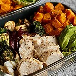 A healthy meal prep container with sliced chicken breast, roasted sweet potatoes, broccoli, onions, and avocado on a kitchen countertop.