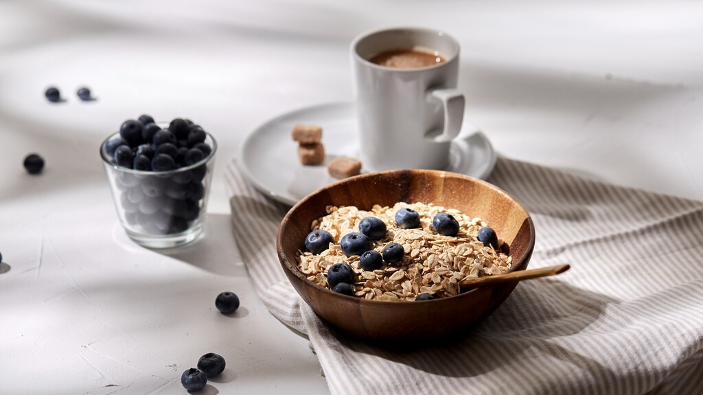 Oatmeal with blueberries: Wooden bowl of oats topped with blueberries, with a spoon, a cup of coffee, and a small bowl of blueberries on a light table.
