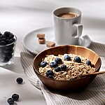 11 Easy Ways to Eat More Fiber (Without Hating Your Meals) Oatmeal with blueberries: Wooden bowl of oats topped with blueberries, with a spoon, a cup of coffee, and a small bowl of blueberries on a light table.