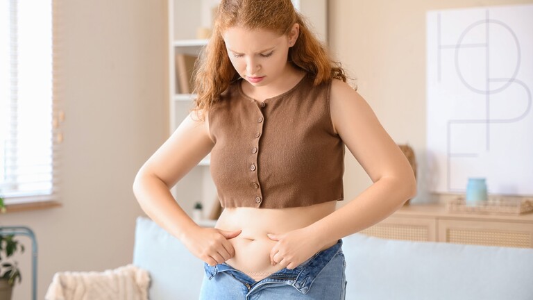 Woman looking down at her midsection while standing in a bright bedroom.