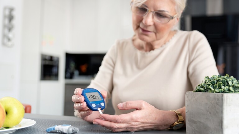 Older woman checking her blood sugar with a glucose meter while sitting at a kitchen table.