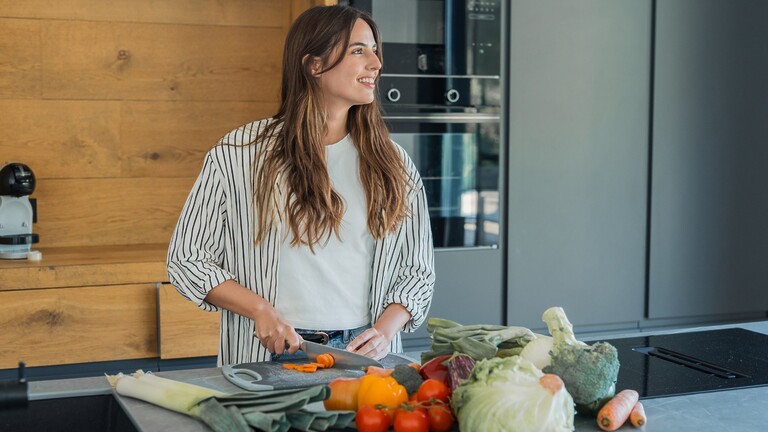 Woman preparing fresh vegetables on a kitchen counter with assorted low-carb produce in the foreground.
