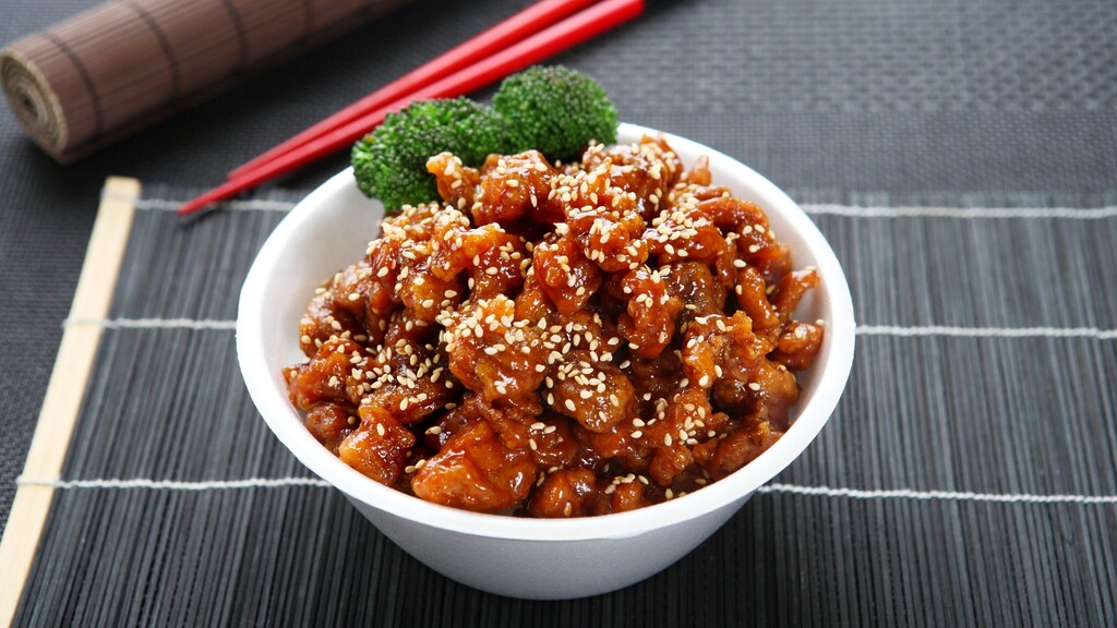 Bowl of sesame chicken with broccoli and red chopsticks on a dark placemat.