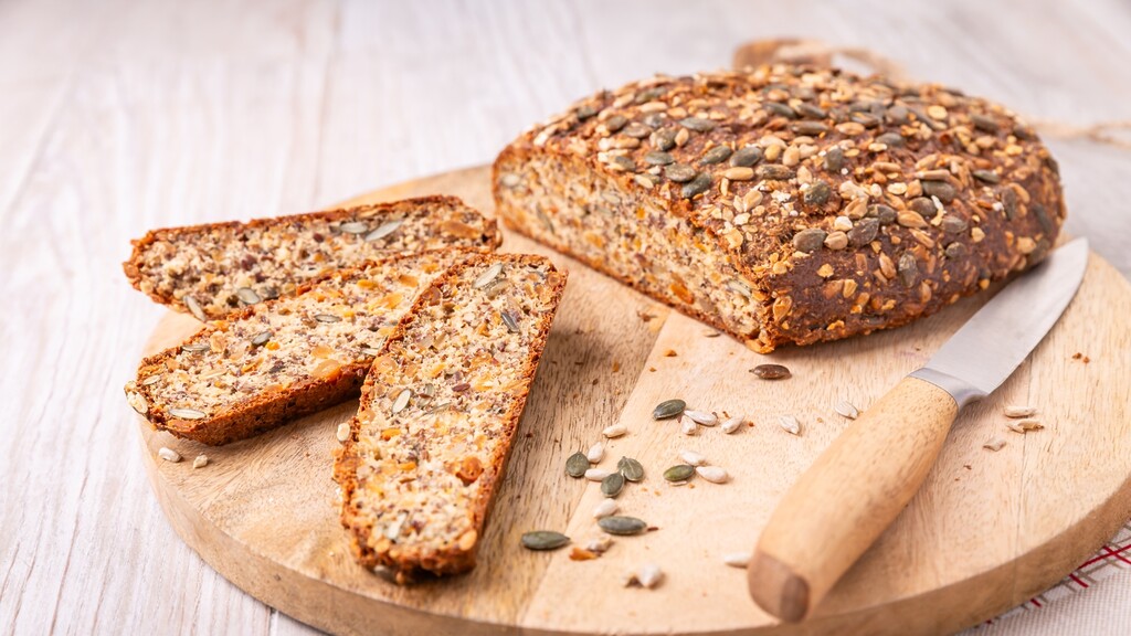 Seeded bread loaf and sliced pieces on a wooden board with a knife beside them.