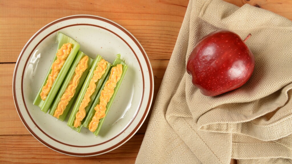 Celery sticks filled with cheese spread served beside a red apple.