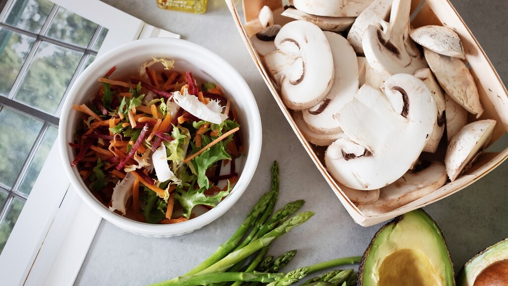 Fresh vegetables on a countertop, including a salad bowl, sliced mushrooms, asparagus, and avocado.