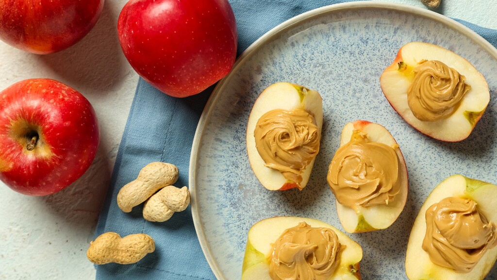 Apple slices topped with peanut butter on a ceramic plate, surrounded by whole red apples and peanuts.