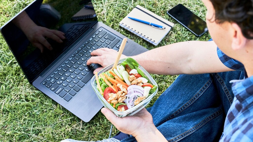 Person working on a laptop while eating a salad outdoors.