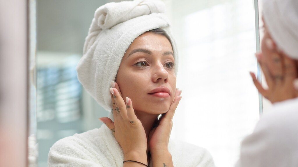 Woman in a white robe and towel looking in a bathroom mirror while touching her face.