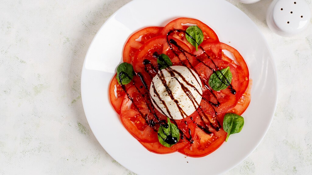 Caprese salad with tomato slices, mozzarella, basil, and balsamic glaze on a white plate.