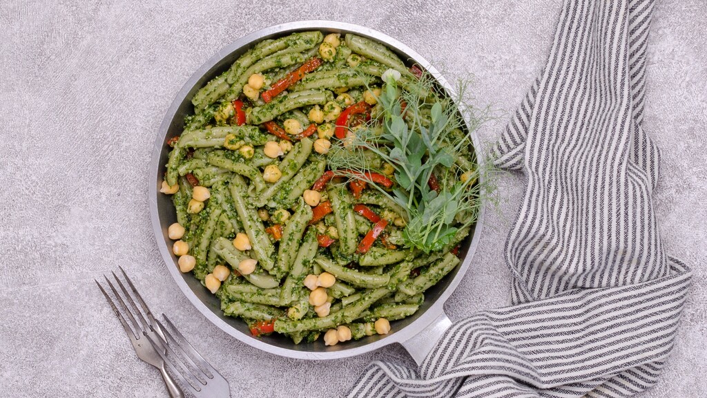Pesto pasta: Bowl of green pesto pasta with chickpeas, red pepper strips, and fresh greens beside a striped cloth.