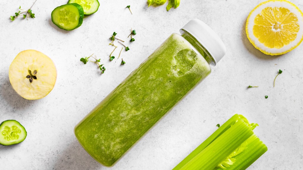 Green juice bottle on a light surface with cucumber slices, celery, apple, lemon, and herbs around it.