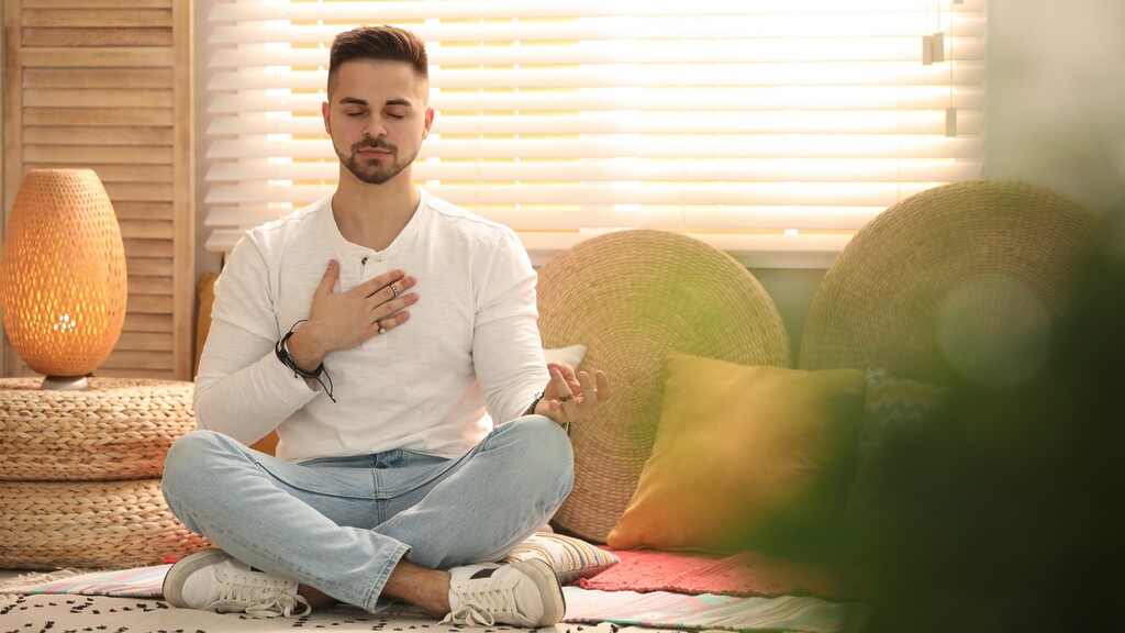 A man sits cross-legged with his eyes closed and one hand on his chest, practicing mindful breathing in a cozy sunlit room.