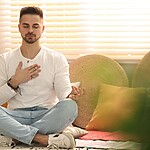 A man sits cross-legged with his eyes closed and one hand on his chest, practicing mindful breathing in a cozy sunlit room.