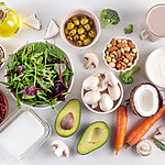 Assorted low-carb foods including leafy greens, avocado, mushrooms, tomatoes, nuts, seeds, olives, coconut, carrots, yogurt, and oil arranged on a white background.