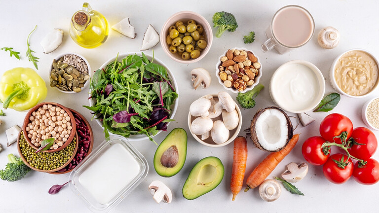 Assorted low-carb foods including leafy greens, avocado, mushrooms, tomatoes, nuts, seeds, olives, coconut, carrots, yogurt, and oil arranged on a white background.