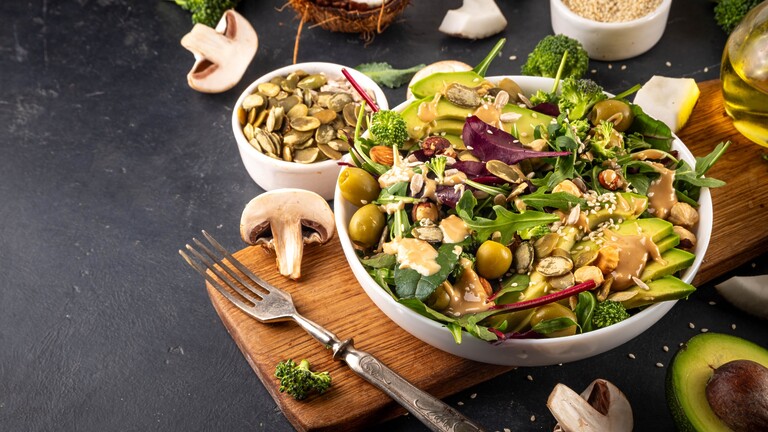 Avocado and mixed green salad with broccoli, olives, seeds, and creamy dressing served in a white bowl on a wooden board.