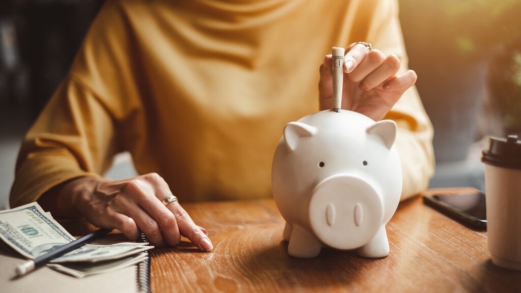 Person placing cash into a white piggy bank on a wooden table, with a notebook, pen, phone, and coffee nearby.