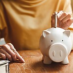 Person placing cash into a white piggy bank on a wooden table, with a notebook, pen, phone, and coffee nearby.
