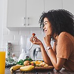 11 Gut Secrets to Stop Hormones From Throwing a Tantrum Woman eating fruit in a bright kitchen with fresh produce on the counter.