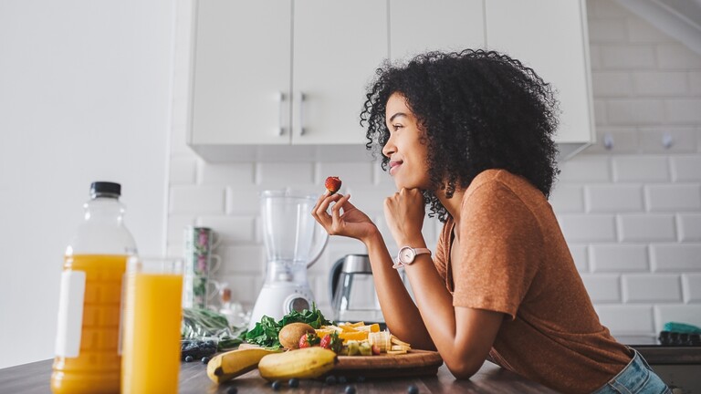 Woman eating fruit in a bright kitchen with fresh produce on the counter.