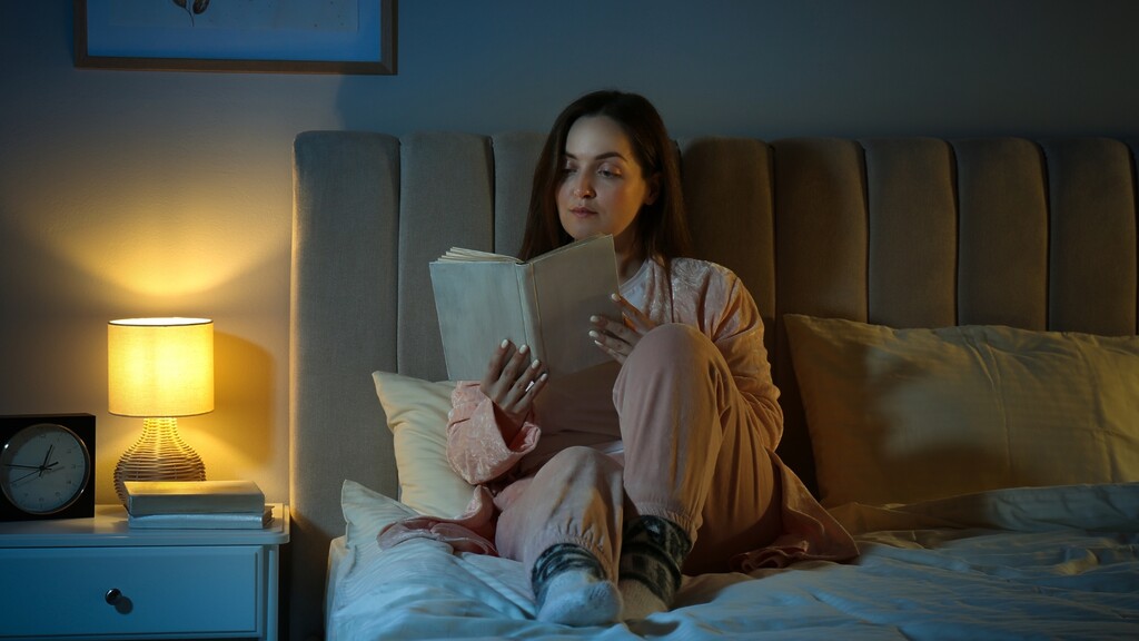 A woman reads in bed beside a warm bedside lamp at night.