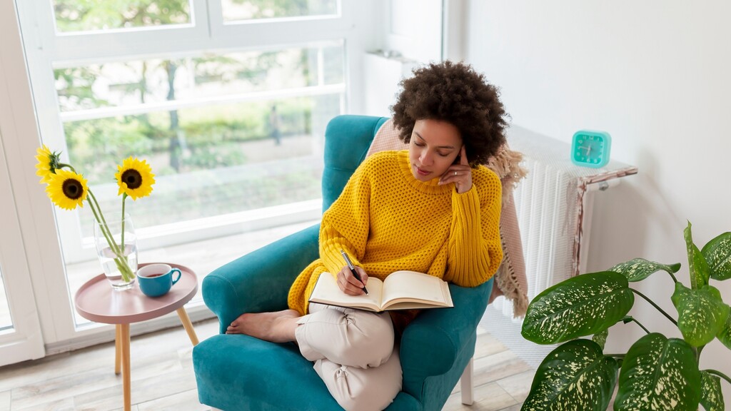 A woman writes in a journal while sitting in a bright armchair near a window.