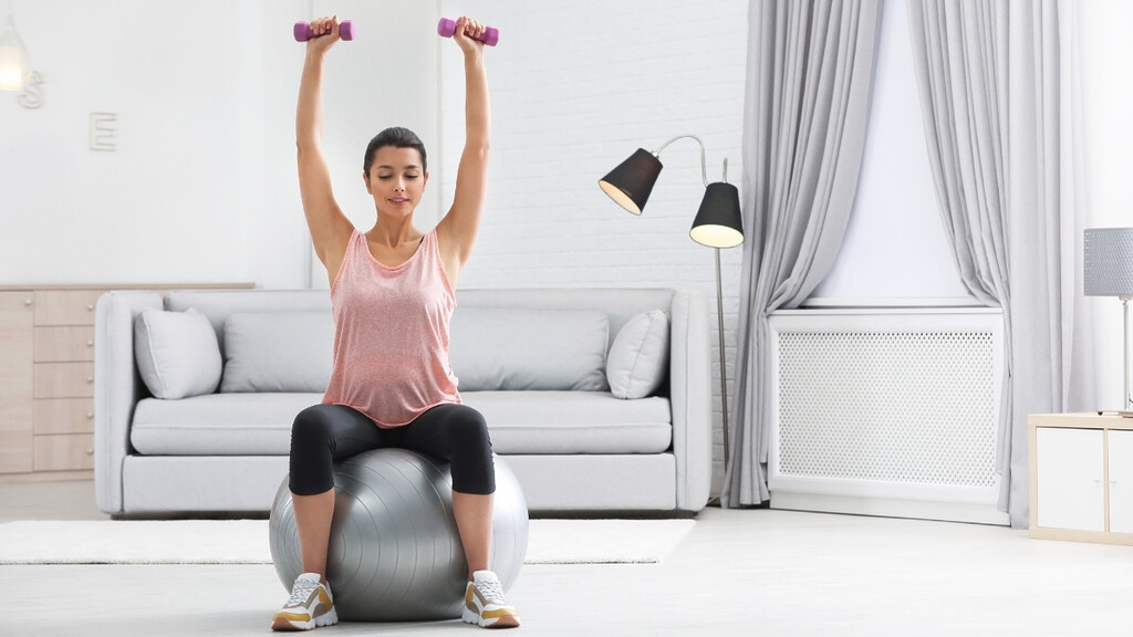 A woman in comfortable workout clothes doing a gentle dumbbell shoulder press in her bright living room.