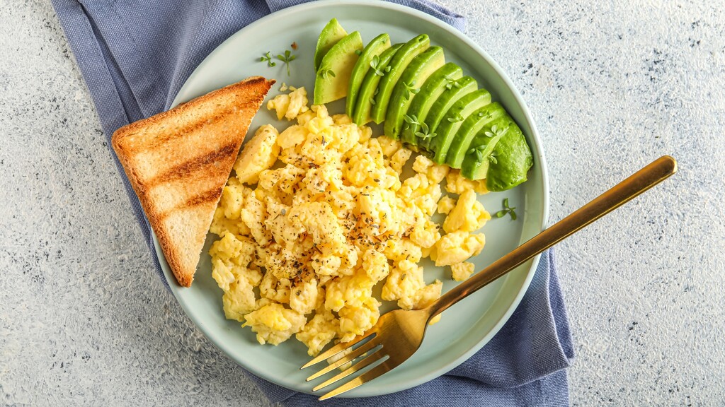 Scrambled eggs with sliced avocado and toast served on a plate with a fork.