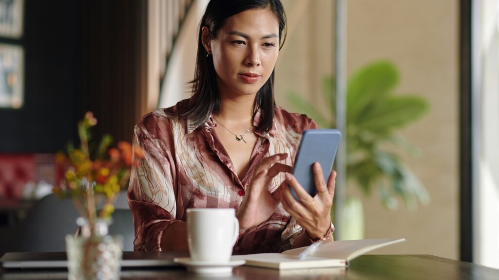 Woman sitting at a table with a coffee and notebook while reviewing her finances on a smartphone.