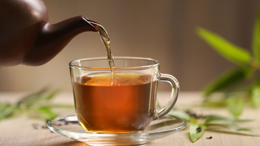 Herbal tea being poured into a clear glass cup beside fresh green leaves.